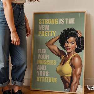 May include: A framed poster featuring a muscular person with dark curly hair wearing a yellow sports bra. The text reads: STRONG IS THE NEW PRETTY, FLEX YOUR MUSCLES AND YOUR ATTITUDE. The background is a muted sage green with light yellow lettering.
