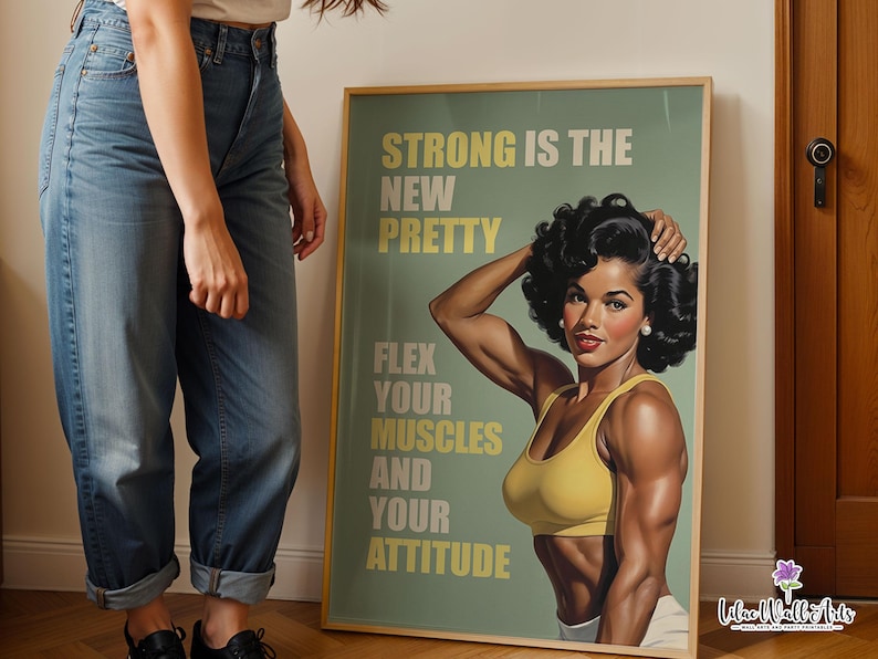 May include: A framed poster featuring a muscular person with dark curly hair wearing a yellow sports bra. The text reads: STRONG IS THE NEW PRETTY, FLEX YOUR MUSCLES AND YOUR ATTITUDE. The background is a muted sage green with light yellow lettering.
