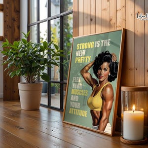 May include: A framed poster featuring a muscular person with dark hair wearing a yellow sports bra, posing with flexed arms. The text reads: STRONG IS THE NEW PRETTY. FLEX YOUR MUSCLES AND YOUR ATTITUDE. The background is a muted olive green color.