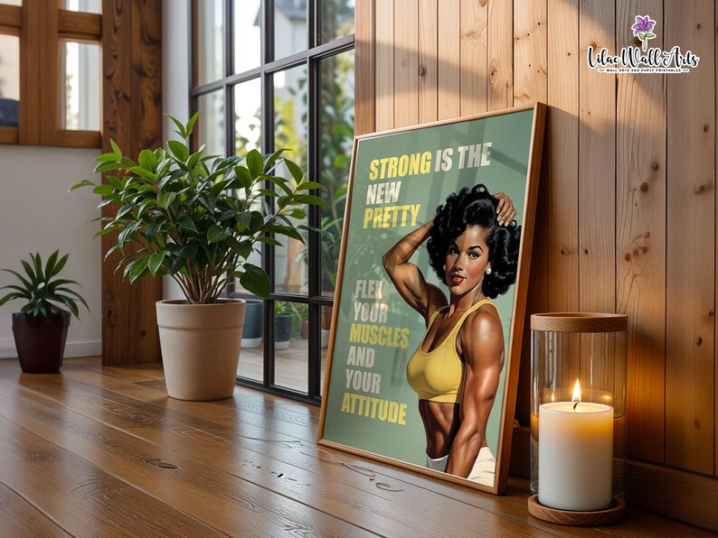 May include: A framed poster featuring a muscular person with dark hair wearing a yellow sports bra, posing with flexed arms. The text reads: STRONG IS THE NEW PRETTY. FLEX YOUR MUSCLES AND YOUR ATTITUDE. The background is a muted olive green color.