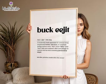 May include: A rectangular wall art print in a light wood frame featuring the black text buck eejit, followed by a phonetic pronunciation, the definition of the Irish slang term, and a list of related words on a white background.