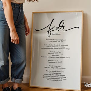 Pode incluir: Uma impressão de arte emoldurada com a palavra "fear" em escrita elegante, acompanhada por um poema de Kahlil Gibran. A impressão tem uma moldura de madeira clara e é exibida contra uma parede neutra.