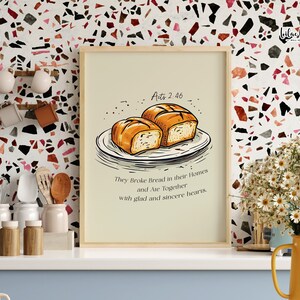 May include: A beige printable wall art with a brown wooden frame. The art features a hand-drawn illustration of two loaves of bread on a plate with the text "Acts 2:46" and "They Broke Bread in their Homes and Ate Together with glad and sincere hearts."