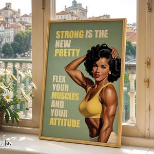 May include: A framed poster featuring a muscular person with dark curly hair wearing a yellow sports bra. The text reads: STRONG IS THE NEW PRETTY. FLEX YOUR MUSCLES AND YOUR ATTITUDE. The background is sage green with yellow lettering.