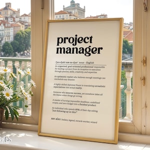 May include: A framed wall art print featuring a dictionary-style definition of the term project manager in bold black text on a cream background. The text describes the role with humour, including phonetic spelling and a see also section at the bottom.