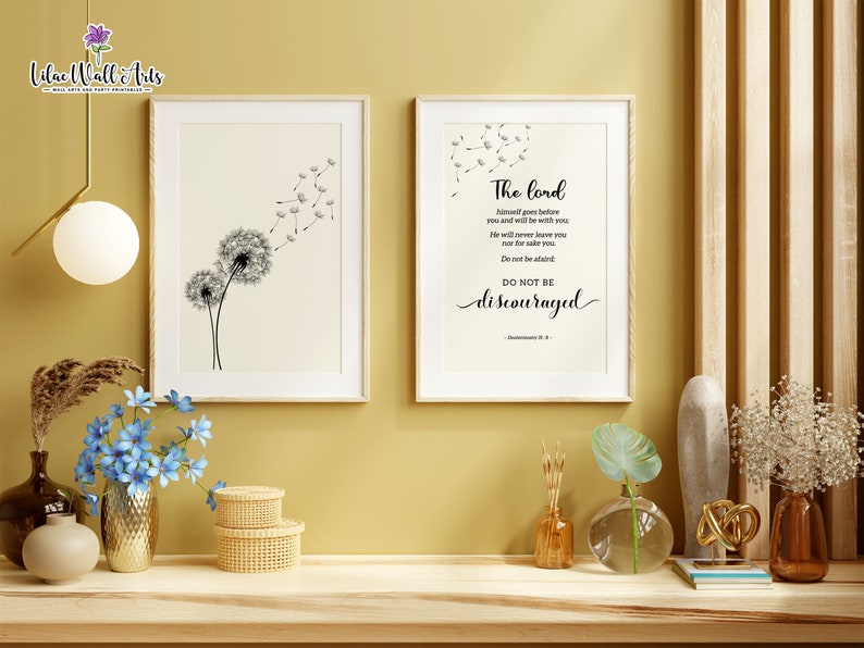 May include: Two framed prints on a wall. The print on the left features a black and white illustration of a dandelion with seeds blowing in the wind. The print on the right features a black and white text quote: "The Lord himself goes before you and will be with you. He will never leave you nor forsake you. Do not be afraid. Do not be discouraged." Deuteronomy 31:8