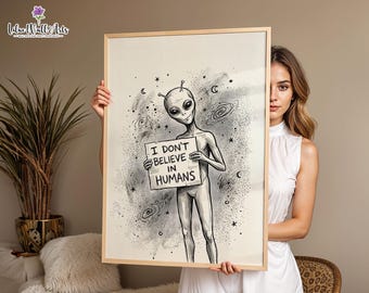 May include: A framed wall art print featuring a black and white illustration of an alien holding a sign that reads I DON'T BELIEVE IN HUMANS. The background includes hand-drawn stars, crescent moons, and spiral galaxies in a sketch style.