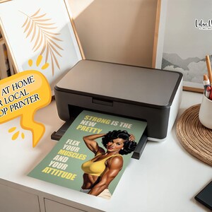 May include: A black and grey desktop printer outputting a printed sheet featuring a muscular individual in a yellow top. The text on the print reads: STRONG IS THE NEW PRETTY, FLEX YOUR MUSCLES AND YOUR ATTITUDE. A yellow speech bubble graphic says PRINT AT HOME OR LOCAL SHOP PRINTER.