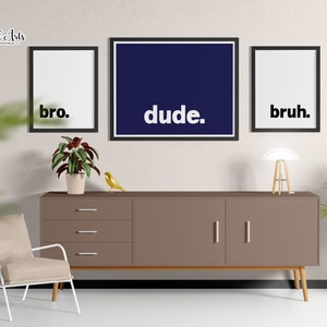 May include: Three framed prints with the words "bro", "dude", and "bruh" in black text on a white background. The prints are hanging on a beige wall above a brown dresser with three drawers and two doors. A small potted plant sits on the dresser, and a lamp is on the right side of the dresser.