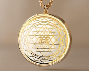 Puede incluir: Collar con colgante dorado con un diseño circular y un patrón geométrico. El colgante tiene un marco dorado y un centro blanco con un diseño triangular detallado. La cadena del collar también es dorada.
