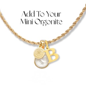 Op de afbeelding: Gouden ketting met een witte bol hanger, een kleine gouden bedel met een geometrisch ontwerp en een gouden letter B bedel. De ketting ligt op een witte achtergrond. Voeg toe aan je mini orgonite.