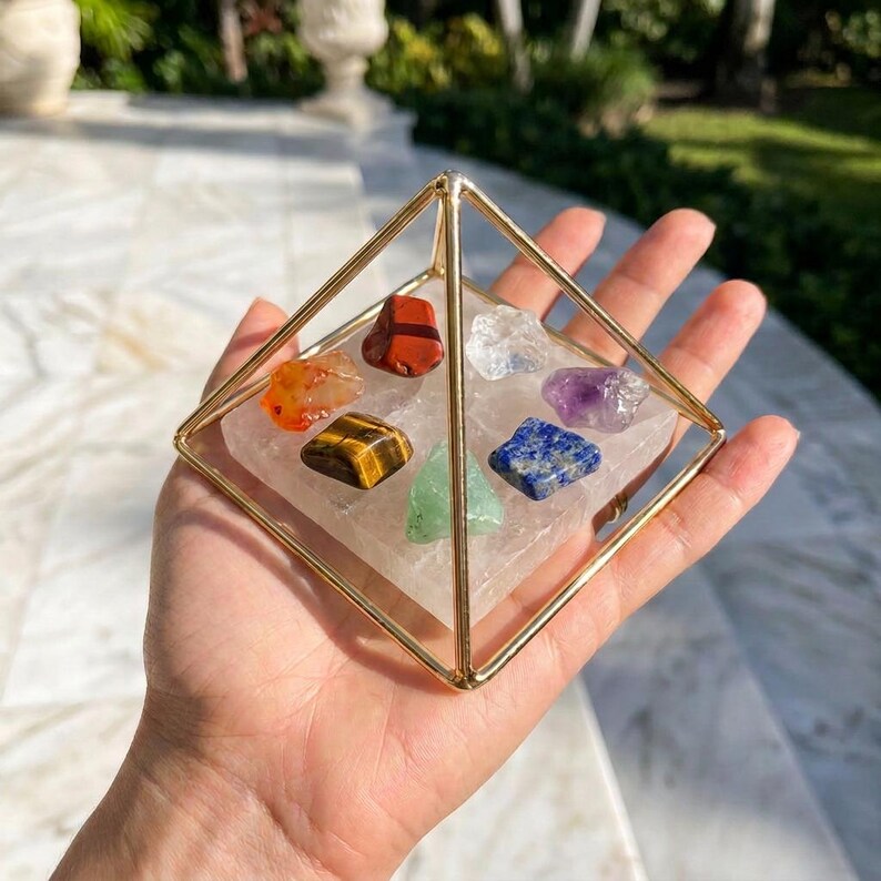 Puede incluir: Una estructura piramidal de metal dorado que encierra una base cuadrada transparente con siete peque&ntilde;as piedras preciosas pulidas y multicolores dispuestas encima, incluyendo variedades en rojo, naranja, amarillo-marr&oacute;n, verde, transparente, azul y morado.