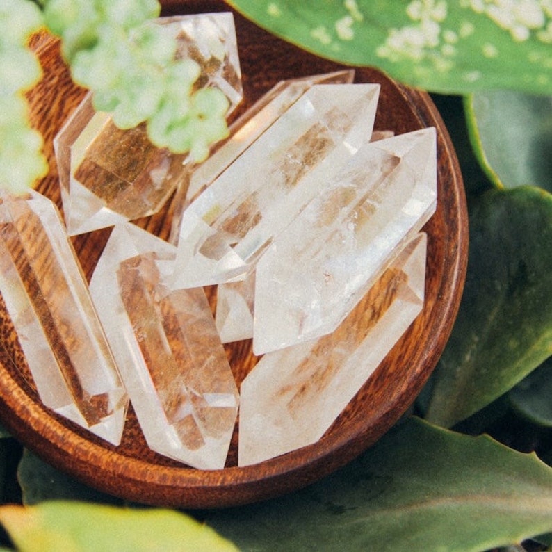 Puede incluir: Una colecci&oacute;n de cristales de cuarzo transparentes de doble terminaci&oacute;n que descansan dentro de un peque&ntilde;o cuenco de madera pulida y redonda. Los cristales son transparentes con facetas internas visibles e inclusiones naturales, dispuestos en grupo.