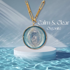 Op de afbeelding: Een goudkleurige ketting met een grote, ronde hanger. De hanger heeft een lichtblauw en wit ontwerp met een centraal kristal. De tekst "Calm & Clear Orgonite" staat in de afbeelding.