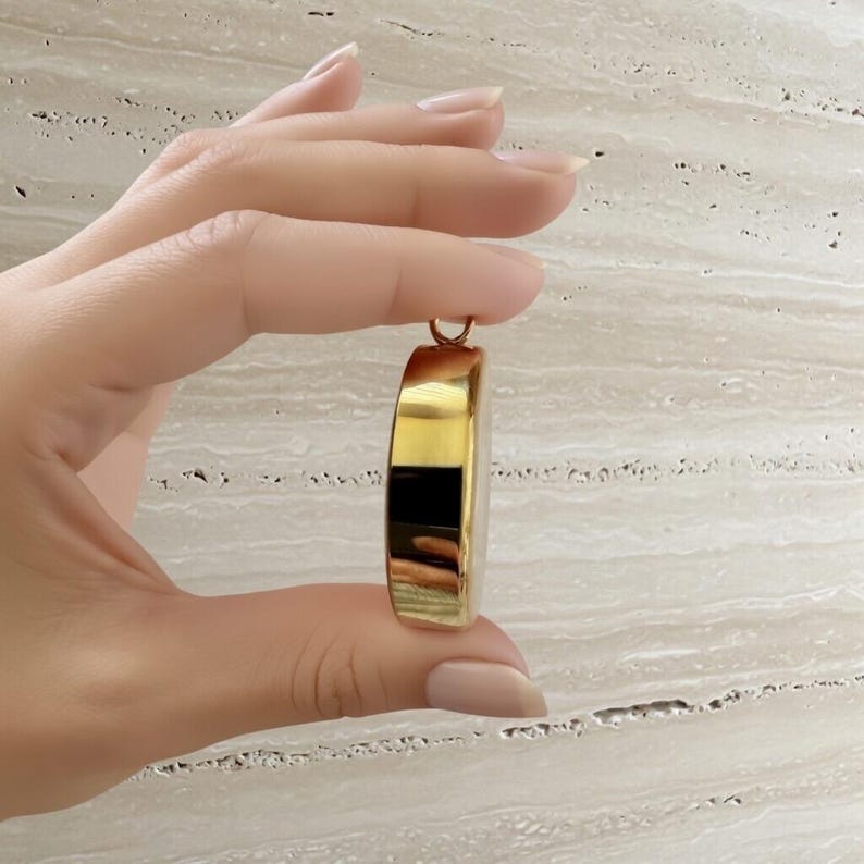May include: A gold-colored pendant with a rounded rectangular shape. The pendant is held by a hand, showcasing its polished surface and design. The pendant has a small loop at the top for a chain.