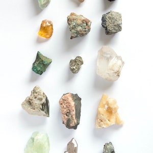 Puede incluir: Una colecci&oacute;n de varios espec&iacute;menes minerales en bruto dispuestos en una cuadr&iacute;cula, que incluye una piedra naranja transl&uacute;cida, un grupo de pirita met&aacute;lica, un cristal de cuarzo transparente, un fragmento de malaquita verde y otras rocas texturizadas en tonos grises, rosas y bronce.