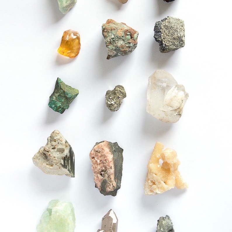 Puede incluir: Una colecci&oacute;n de varios espec&iacute;menes minerales en bruto dispuestos en una cuadr&iacute;cula, que incluye una piedra naranja transl&uacute;cida, un grupo de pirita met&aacute;lica, un cristal de cuarzo transparente, un fragmento de malaquita verde y otras rocas texturizadas en tonos grises, rosas y bronce.