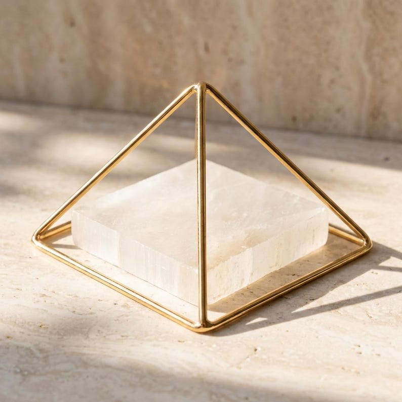 Puede incluir: Un marco de alambre met&aacute;lico de color dorado con forma de estructura piramidal. Dentro de la base de la pir&aacute;mide de alambre dorado se asienta una losa de piedra cuadrada, blanca y transl&uacute;cida, con una superficie lisa y aspecto cristalino.
