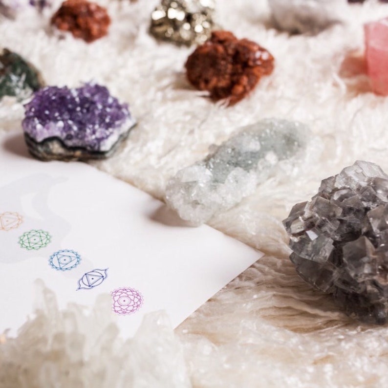 Puede incluir: Una colecci&oacute;n de varios cristales minerales en bruto, incluyendo un racimo de amatista p&uacute;rpura, una pieza de pirita met&aacute;lica y una formaci&oacute;n de cristal gris oscuro, dispuestos junto a una tarjeta blanca con cinco s&iacute;mbolos de chakra circulares coloridos.