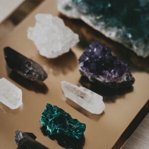 Puede incluir: Una colecci&oacute;n de espec&iacute;menes minerales en bruto que incluye un grupo de cuarzo blanco, una drusa de amatista de color p&uacute;rpura intenso, una punta de cuarzo ahumado, un cristal de cuarzo transparente, una pieza de malaquita verde y un cristal gris oscuro sobre una bandeja dorada.