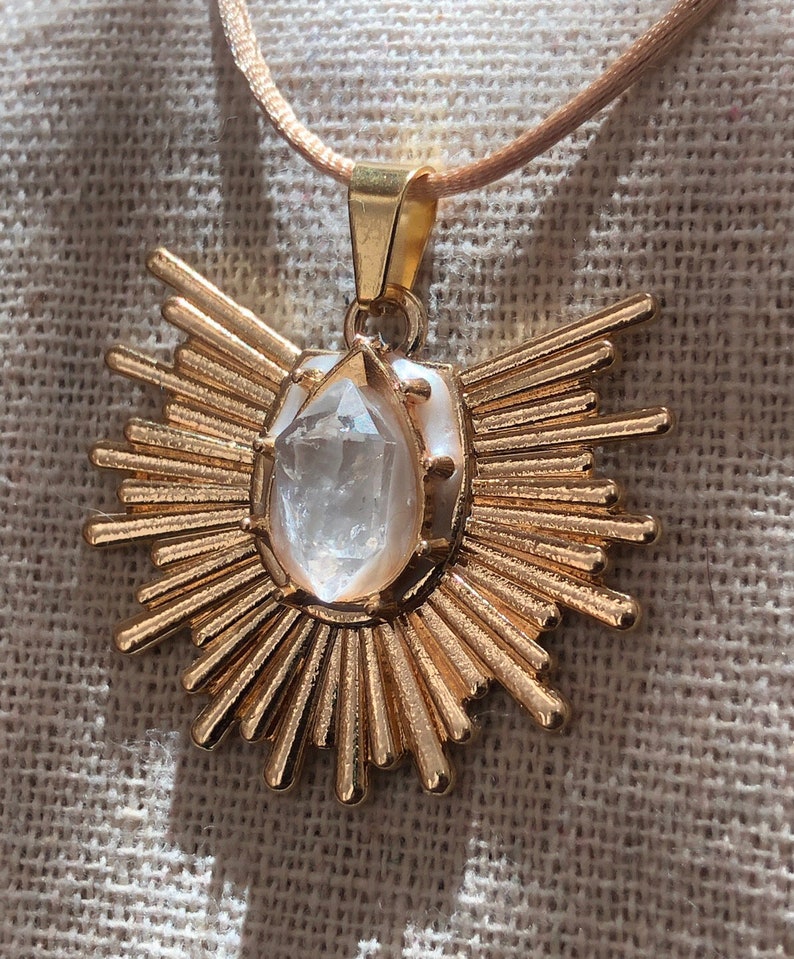 May include: A gold pendant necklace featuring a clear quartz crystal at its centre. The pendant is shaped like a sun with numerous rays.