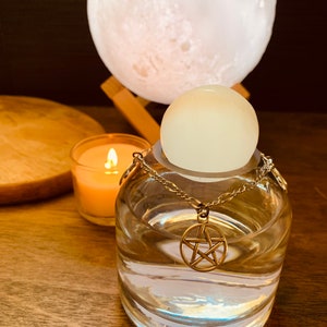 Peut inclure: Une bouteille en verre transparente avec une chaîne argentée et un pendentif pentagramme à l'intérieur. La bouteille est posée sur une surface en bois avec une lampe lunaire blanche et une bougie en arrière-plan.