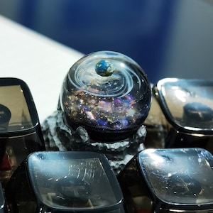 Könnte beinhalten: Eine transparente Harz-Tastenkappe mit einem Galaxie-Design und einem kleinen blauen Planeten in der Mitte. Die Tastenkappe ist von anderen schwarzen Tastenkappen mit einem Kreuz-Design umgeben.