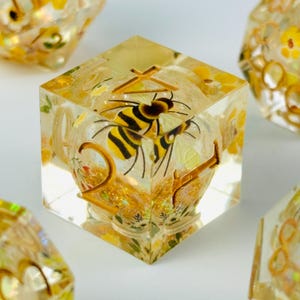 May include: A set of dice with a bee theme. The dice are clear with yellow and gold accents, and a bee is embedded inside each die. The numbers are gold. The dice are surrounded by other dice with similar designs.