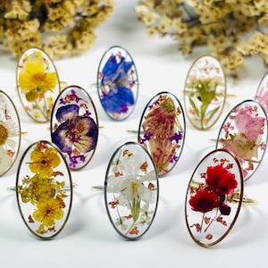 Puede incluir: Un conjunto de anillos ovalados con engastes de resina transparente, cada uno con una flor seca diferente. Las flores son de varios colores, incluyendo amarillo, morado, blanco y rojo, engastadas en bandas plateadas o doradas. Los anillos se exhiben sobre una superficie blanca.