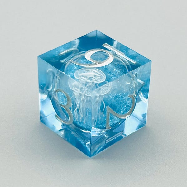 May include: A clear blue dice with a white jellyfish inside. The dice has the numbers 1, 2, 3, 4, 5, and 6 on its sides.