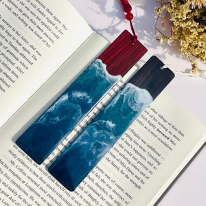 May include: Two handmade resin bookmarks with a wave design. The bookmarks are on a book with text. One bookmark is blue and white, the other is green and white. Both bookmarks have a wooden base.