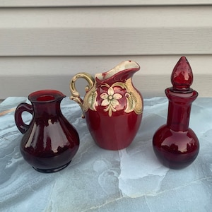 May include: Three red glass bottles, one with a floral design, one with a round body, and one with a tall, slender body. The bottles are all in a vintage style.
