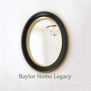 May include: An oval wall mirror featuring a matte black outer frame and a thin gold inner rim. The reflective glass surface shows a partial reflection of a crystal chandelier and white curtains. The text Baylor Home Legacy appears below.