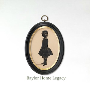May include: An oval wall frame with a black outer border and a thin gold inner rim, featuring a silhouette portrait of a person in a hat and coat on a cream background. A small metal hanging loop is attached to the top. Text reads Baylor Home Legacy.