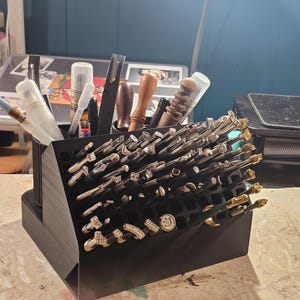 May include: A black organizer filled with various jewelry-making tools and stamps. The organizer is angled, displaying the tools for easy access. A variety of tools are visible, including pens, brushes, and stamps with intricate designs. The background includes a desk and a lamp.