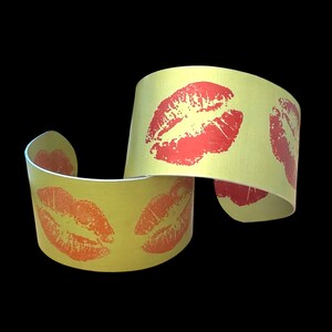 May include: A yellow cuff bracelet featuring multiple red lip prints. The bracelet is made of a flexible material with a metallic finish. The lip prints are evenly spaced around the bracelet. The design is bold and eye-catching.