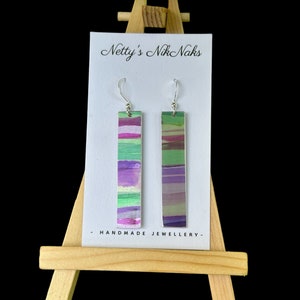 May include: A pair of handmade dangle earrings with a silver hook and a rectangular green, purple, and white striped design. The earrings are displayed on a white card with the text "Netty's Nik Naks - HANDMADE JEWELLERY - ".