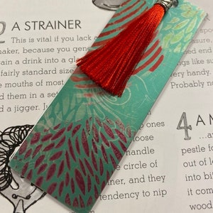 Könnte beinhalten: Ein türkisfarbenes Lesezeichen mit einer roten Quaste. Das Lesezeichen hat ein florales Design in Rot und Grün. Die Quaste besteht aus roten Fäden und ist mit einer silberfarbenen Metallkappe befestigt.