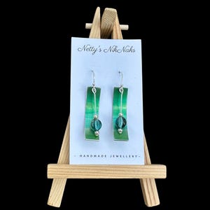 Puede incluir: Pendientes hechos a mano exhibidos en un pequeño caballete de madera. Los pendientes presentan un diseño rectangular verde y plateado con una cuenta turquesa colgante y detalles plateados. La tarjeta dice "Netty's Nik Naks - HANDMADE JEWELLERY-".