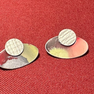 May include: A pair of oval-shaped earrings with a textured silver circle on top of each. The earrings have a red, yellow, and grey paint design.