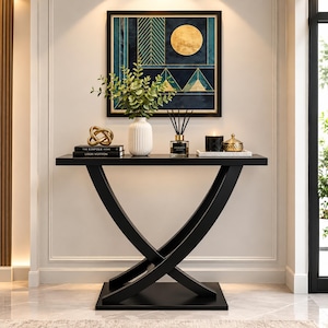 May include: A black console table with a curved, intersecting base and a rectangular top. On the table are two stacked books, a gold knot sculpture, a white ribbed vase with green foliage, a reed diffuser, and a black candle with a gold lid.