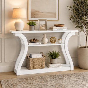 May include: A white console table with a curved frame design, featuring two open shelves. The table holds a lamp, books, a woven basket, potted plants, a picture frame, a decorative bowl, and a gold geometric object on its surfaces.