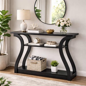 May include: A black console table with a rectangular top, a middle shelf, and a bottom shelf, featuring curved side supports. The table holds a white lamp, a coral sculpture, books, a bowl, a clock, a vase of white flowers, a woven basket, and a plant.
