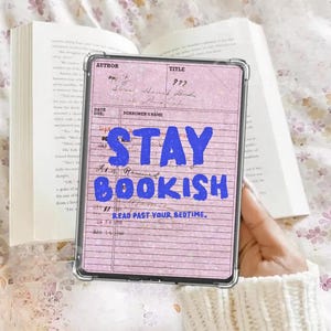 Può includere: Una custodia trasparente per tablet con sfondo rosa con le parole "STAY BOOKISH" in blu. Il tablet è tenuto davanti a un libro aperto. È visibile anche il testo "READ PAST YOUR BEDTIME".