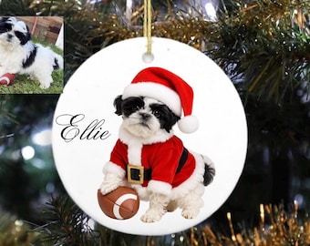 Puede incluir: Adorno navideño redondo con un cachorro Shih Tzu vestido con un traje de Papá Noel, que incluye un gorro y un traje rojos. El cachorro sostiene un balón de fútbol americano. El nombre "Ellie" está escrito en cursiva. El adorno cuelga de una cuerda dorada.