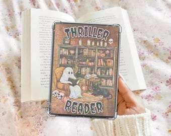 May include: A clear phone case featuring a spooky illustration of a bookshelf, ghost, and black cat, with the words "THRILLER READER". The case is held over an open book on a floral patterned surface.