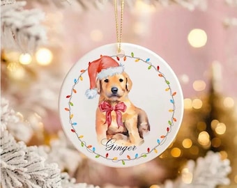 May include: A circular Christmas tree decoration with a golden retriever puppy wearing a Santa hat and a red bow. The puppy's name, "Ginger," is written in cursive. The ornament is encircled by a string of colourful lights.
