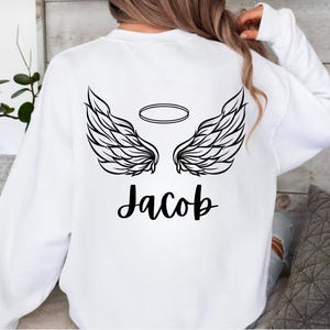 May include: White sweatshirt featuring a black graphic of angel wings and a halo above the name "Jacob". The design is centered on the back of the sweatshirt.