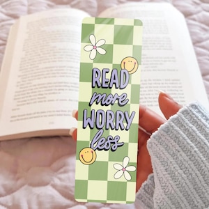 Könnte beinhalten: Ein rechteckiger Lesezeichen mit einem grün-weißen Karomuster. Das Lesezeichen zeigt den Text "READ more WORRY less" in einer verspielten Schriftart, zusammen mit Smiley-Gesichtern und Blumenillustrationen. Das Lesezeichen wird vor einem offenen Buch gehalten.