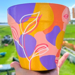 May include: A hand-painted terracotta plant pot featuring an abstract pattern of overlapping organic shapes in vibrant shades of yellow, purple, orange, and light pink, accented with thin, white, hand-drawn line art of leaves and stems.